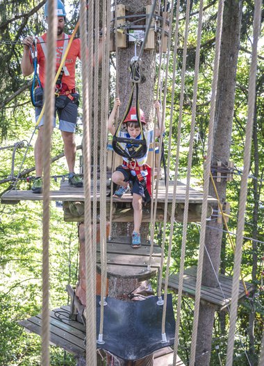 Kinder beim Kletterspaß im Klettergarten Söllereck, umgeben von Bäumen, mit Sicherheitsausrüstung, unter Anleitung eines Erwachsenen. | © Oberstdorf · Kleinwalsertal Bergbahnen