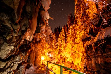 Die Breitachklamm im Dunkeln bei Nacht, beleuchtet von warmen Lichtern, mit schroffen Felsen und verschneiten Bäumen. | © Kleinwalsertal Tourismus eGen • Breitachklamm • Fotograf: Domink Berchtold