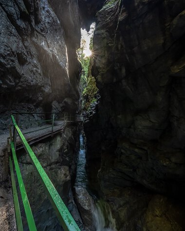 Blick durch die Breitachklamm im Sommer mit Wasserfall und schroffen Felswänden, Sonne am Himmel sichtbar. | © Kleinwalsertal Tourismus eGen • Breitachklamm • Fotograf: Dominik Berchtold