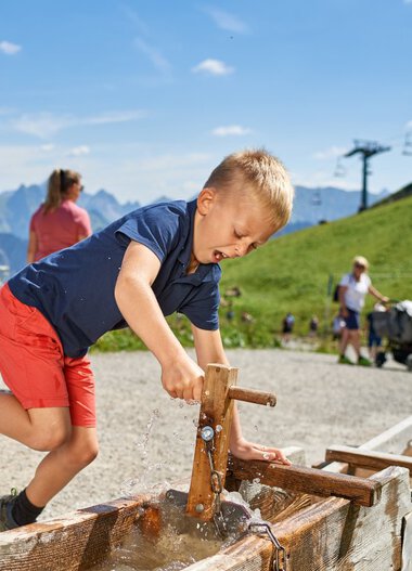Junge spielt mit Wasserhahn im Freien in den Bergen, springt vor Freude. | © Oberstdorf · Kleinwalsertal Bergbahnen Fotograf: Christian Seitz