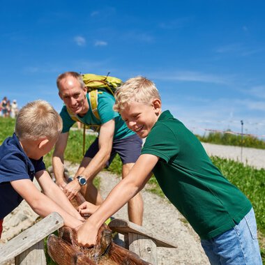 Familie mit zwei Jungen trinkt frisches Wasser aus einer natürlichen Quelle im Grünen an einem sonnigen Tag in Oberstdorf. | © Oberstdorf · Kleinwalsertal Bergbahnen Fotograf: Christian Seitz