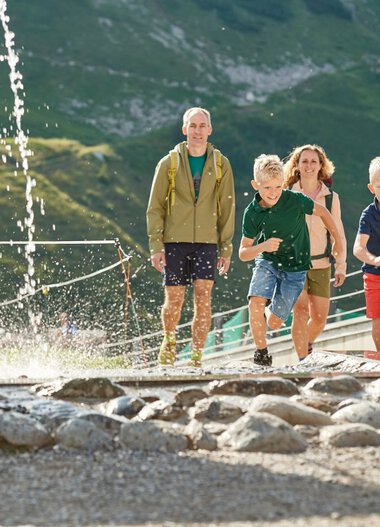 Familie mit zwei Kindern läuft barfuß durch einen Wasserlauf in den Bergen, im Hintergrund grüner Bergwald und eine Seilbahn. | © Oberstdorf · Kleinwalsertal Bergbahnen Fotograf: Christian Seitz