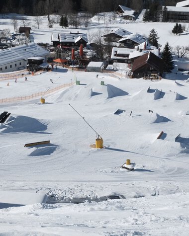 Snowpark am Crystal Ground mit verschiedenen Sprüngen und Hindernissen im Schnee, umgeben von einer verschneiten Berglandschaft und Gebäuden. | © Kleinwalsertal Tourismus eGen • Crystal Ground
