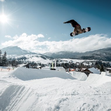 Snowboarder fliegt bei Sonnenlicht über eine Snowpark-Sprungrampe in einer verschneiten Winterszenerie. | © Kleinwalsertal Tourismus eGen • Crystal Ground • Fotograf: Stefan Eigner