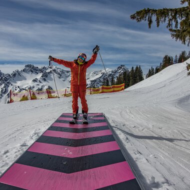 Skifahrer in roter Kleidung auf einer bestimmten Skipiste in Oberstdorf, mit verschneiten Bergen im Hintergrund und einem blauen Himmel. | © Kleinwalsertal Tourismus eGen • Oberstdorf Kleinwalsertal Bergbahnen