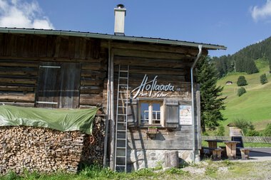 Traditionelle Holzhütte mit Holzlager und Aufstiegslatte, inmitten grüner Berge, Werbung für den Hofladen im Kleinwalsertal. | © Kleinwalsertal Tourismus eGen • Hoflaada