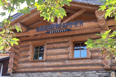 Holzhaus mit zwei kleinen Fenstern, das wahrscheinlich eine Rauchkammer ist, umgeben von grünen Blättern. | © Kleinwalsertal Tourismus eGen • Räucherkammer