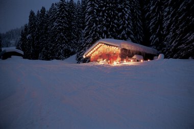 Ein verschneites Holzhaus mit Weihnachtslichtern in einer winterlichen, dunklen Landschaft, umgeben von hohen Tannenbäumen. | © Kleinwalsertal Tourismus eGen • Max Hütte • Fotograf: Hans Wiesenhofer