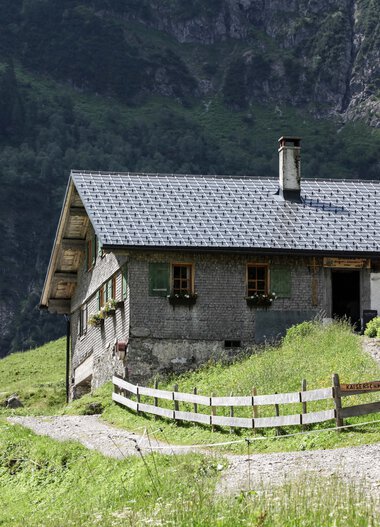 Bernhards Gemstelhütte in den Bergen, umgeben von grüner Natur und Wald, mit Terrasse und Schirm. Dach aus Schindeln, kleiner Weg leading zur Hütte. | © Kleinwalsertal Tourismus eGen • Bernhards Gemstelhütte • Fotograf: Frank Drechsel