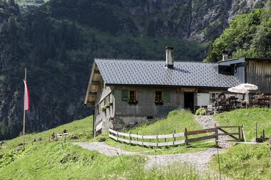 Bernhards Gemstelhütte in den Bergen, umgeben von grüner Natur und Wald, mit Terrasse und Schirm. Dach aus Schindeln, kleiner Weg leading zur Hütte. | © Kleinwalsertal Tourismus eGen • Bernhards Gemstelhütte • Fotograf: Frank Drechsel