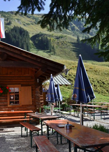 Bergrestaurant in den Alpen mit mehreren Tischen und schwarzen Sonnenschirmen im Außenbereich, umgeben von grünen Wiesen und Bergen. | © Kleinwalsertal Tourismus eGen • Mittelalp