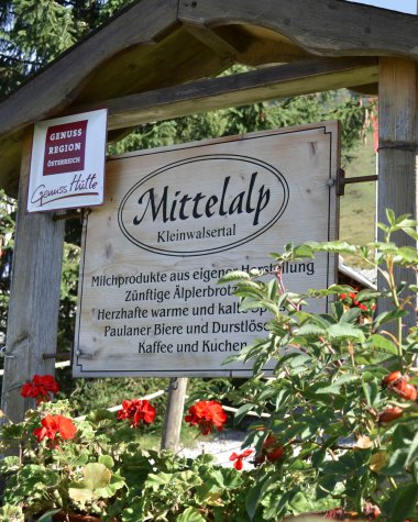Hinweisschild für die GenussRegion Mittelalp in Kleinwalsertal, umgeben von roten Blumen und grünen Pflanzen. Im Hintergrund Holzpfosten und Bäume. | © Kleinwalsertal Tourismus eGen • Mittelalp