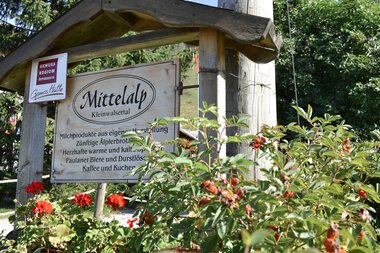 Hinweisschild für die GenussRegion Mittelalp in Kleinwalsertal, umgeben von roten Blumen und grünen Pflanzen. Im Hintergrund Holzpfosten und Bäume. | © Kleinwalsertal Tourismus eGen • Mittelalp