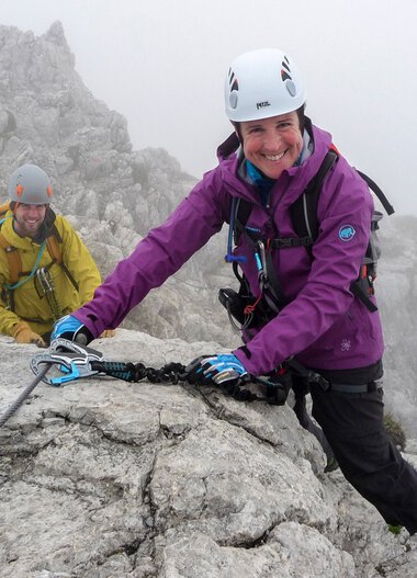 Zwei Kletterer am Hindelanger Klettersteig, eine lächelnde Frau in lila Jacke, im Nebel auf einer felsigen Bergwand. | © Kleinwalsertal Tourismus eGen • Alpinschule Oberstdorf