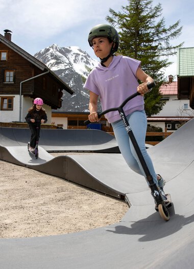 Zwei Kinder, ein Mädchen und ein Junge, fahren auf Scootern auf einer Skate-Anlage in den Bergen mit Holz- und Steinhäusern im Hintergrund. | © Kleinwalsertal Tourismus eGen • Fotograf: Frank Drechsel