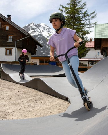 Zwei Kinder, ein Mädchen und ein Junge, fahren auf Scootern auf einer Skate-Anlage in den Bergen mit Holz- und Steinhäusern im Hintergrund. | © Kleinwalsertal Tourismus eGen • Fotograf: Frank Drechsel