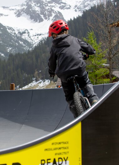 Zwei Kinder fahren mit Fahrrädern auf einem Pumptrack in einer bergigen Landschaft mit Schnee bedeckten Bergen im Hintergrund. | © Kleinwalsertal Tourismus eGen • Fotograf: Frank Drechsel