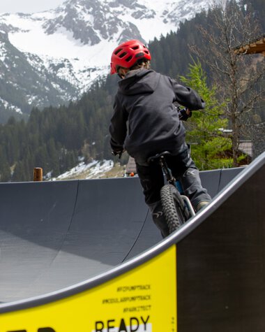 Zwei Kinder fahren mit Fahrrädern auf einem Pumptrack in einer bergigen Landschaft mit Schnee bedeckten Bergen im Hintergrund. | © Kleinwalsertal Tourismus eGen • Fotograf: Frank Drechsel
