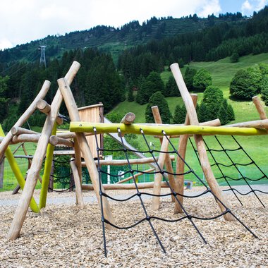 Spielplatz mit Klettergerüst aus Holz und Seilen im Grünen, im Hintergrund Berge und Bäume, am Mittelberg. | © Kleinwalsertal Tourismus eGen