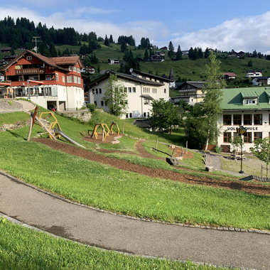 Blick auf einen Spielplatz in Hirschegg mit Rutsche, Schaukeln und Wegen, umgeben von grüner Wiese und Bergen im Hintergrund. | © Kleinwalsertal Tourismus eGen