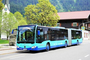 Ein blauer Bus steht an einer Haltestelle in einer ländlichen Gegend, umgeben von Bäumen und Häusern. | © Kleinwalsertal Tourismus eGen