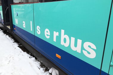 Nahaufnahme eines Walsebus im Winter mit Schnee an den Rädern und auf dem Boden, Seitenschriftzug 'Walserbus' in weißen Buchstaben auf türkisblauer Hintergrund. | © Kleinwalsertal Tourismus eGen