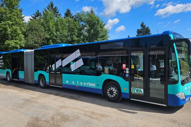 Moderner Wasserbus in Blau mit schwarzem und weißen Design, geparkt auf einer Straße mit Bäumen und blauem Himmel im Hintergrund. | © Kleinwalsertal Tourismus eGen