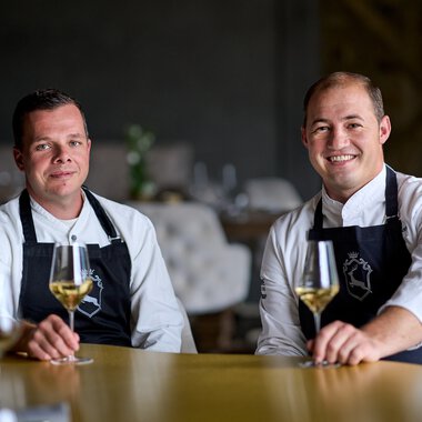 Zwei professionelle Köche sitzen mit Weingläsern in einem eleganten Restaurant, bereit für ein kulinarisches Erlebnis. | © Kleinwalsertal Tourismus eGen • Anette Sandner