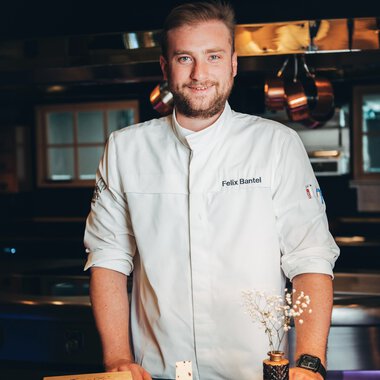 Ein männlicher Koch in weißer Approbation steht hinter einer Theke in einer professionellen Küche, mit einem kleinen dekorativen Dessert in der Hand. | ©  Kleinwalsertal Tourismus eGen • Hotel Birkenhöhe
