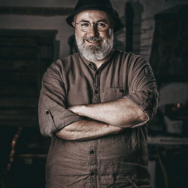 Ein Mann mit Brille und Bart trägt einen schwarzen Hut und ein braunes Hemd, steht vor einer rustikalen Holzfassade, lächelt und hat die Arme verschränkt. | © Kleinwalsertal Tourismus eGen • Biohotel Walserstuba • Fotograf: Moving Stills