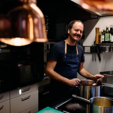 Ein fröhlicher Koch in einer modernen Küche, beim Kochen mit einem großen Topf, umgeben von Küchenutensilien und Getränkeflaschen an der Wand. | © Kleinwalsertal Tourismus eGen • Fotograf: Oliver Farys