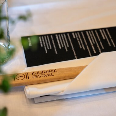Menükarte für das Kulinarikfestival 2025 auf einem Tisch, mit einem Smartphone, Papier und einem Glas | © Kleinwalsertal Tourismus eGen • Fotograf: Isenhoff