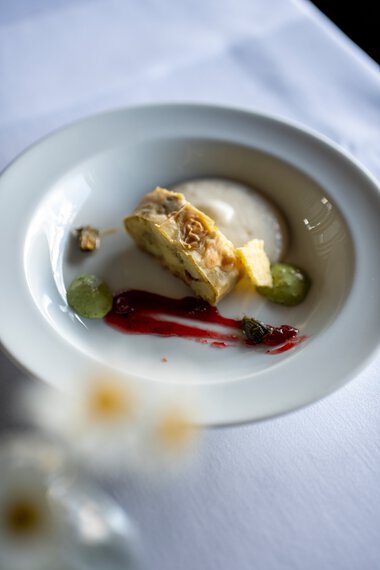 Feine Portion einer modernen Dekorations- oder Gourmet-Speise auf einem weißen Teller, mit cremiger Soße, Gemüsescheiben und einer roten Balsamico-Reduktion. | © Kleinwalsertal Tourismus eGen • Fotograf: Isenhoff