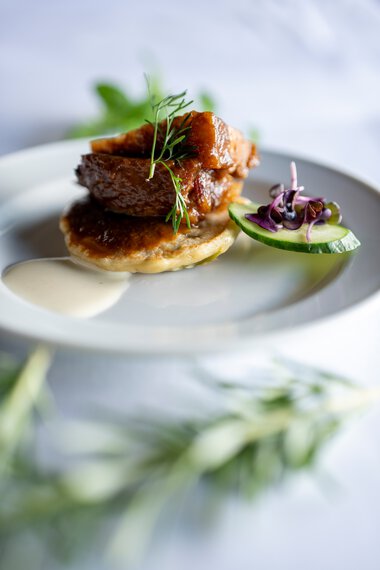 Elegantes Gericht mit knusprigem Fleisch auf cremigem Grund, garniert mit frischer Kräutergarnitur und eingelegtem Gemüse, serviert auf einem weißen Teller. | © Kleinwalsertal Tourismus eGen • Fotograf: Isenhoff