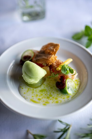Elegantes Gericht mit grünem Sorbet, frittiertem Stück Fleisch, verschiedenen Gemüsen und einer leichten Sauce, serviert auf weißem Teller. | © Kleinwalsertal Tourismus eGen • Fotograf: Isenhoff