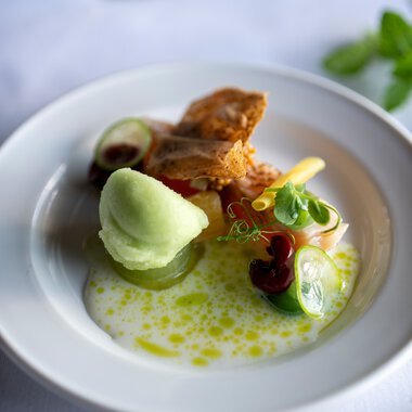 Elegantes Gericht mit grünem Sorbet, frittiertem Stück Fleisch, verschiedenen Gemüsen und einer leichten Sauce, serviert auf weißem Teller. | © Kleinwalsertal Tourismus eGen • Fotograf: Isenhoff