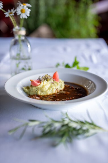 Ein Teller mit Kartoffelstampf und brauner Soße, dekoriert mit kleinen Tomaten und Blüten, auf einem weißen Tischtuch bei einem Kulinarikfestival. | © Kleinwalsertal Tourismus eGen • Fotograf: Isenhoff