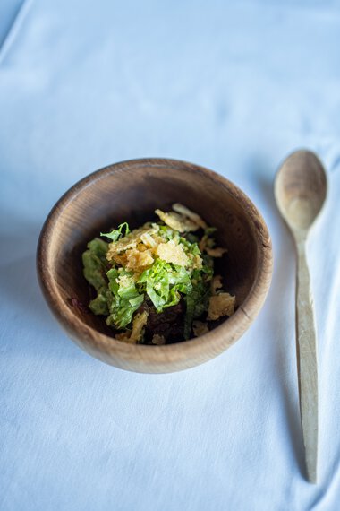Ein kleiner Holztopf mit frisch zubereitetem Salat auf weißem Tischtuch, daneben ein Holzlöffel. | © Kleinwalsertal Tourismus eGen • Fotograf: Isenhoff