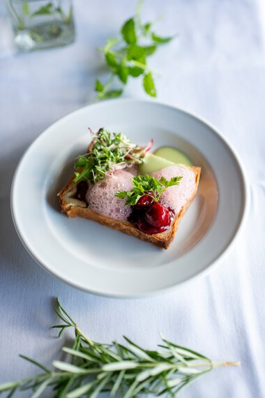 Elegantes Gericht auf einem weißen Teller: ein Toast mit Fleisch, roten Beeren, grüner Soße und Salat, dekoriert mit frischen Kräutern. | © Kleinwalsertal Tourismus eGen • Fotograf: Isenhoff
