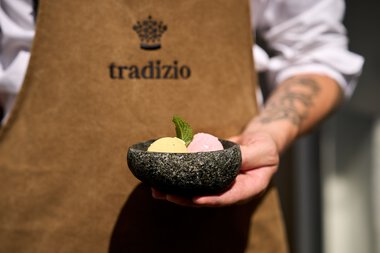 Person hält eine Stein-Schale mit zwei Kugeln Eis und Minzblatt, im Hintergrund ein Tragebald mit 'tradizio' Logo. | © Kleinwalsertal Tourismus eGen • Hotel Tradizio • Fotograf: Annette Sandner