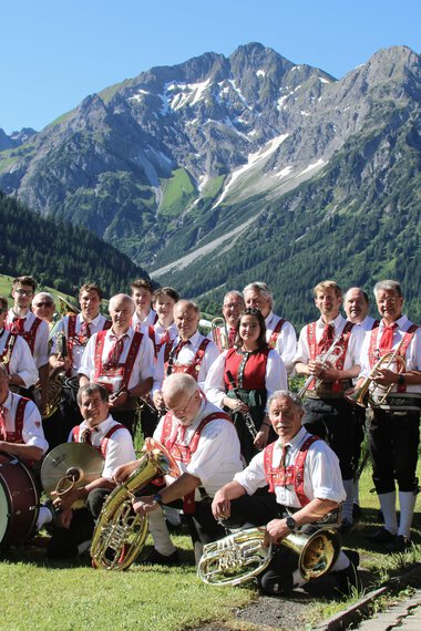 Musikkapelle in traditioneller Tracht vor einer Bergkulisse mit hohen Bergen und Wäldern, Gruppe mit Instrumenten und Banner. | © Kleinwalsertal Tourismus eGen • Musikverein d'Hirschegger