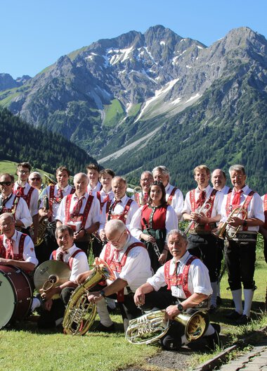 Musikkapelle in traditioneller Tracht vor einer Bergkulisse mit hohen Bergen und Wäldern, Gruppe mit Instrumenten und Banner. | © Kleinwalsertal Tourismus eGen • Musikverein d'Hirschegger