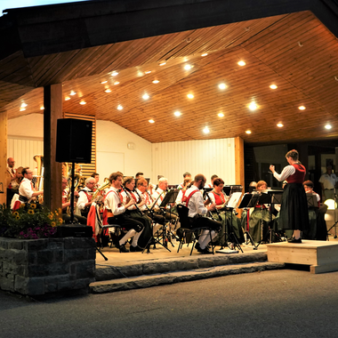 Musikkapelle spielt auf einer Bühne im Freien bei Nacht, beleuchtet von warmen Lichtern, mit Dirigentin und Musikern in traditionellen Trachten. | ©  Kleinwalsertal Tourismus eGen