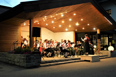 Musikkapelle spielt auf einer Bühne im Freien bei Nacht, beleuchtet von warmen Lichtern, mit Dirigentin und Musikern in traditionellen Trachten. | ©  Kleinwalsertal Tourismus eGen