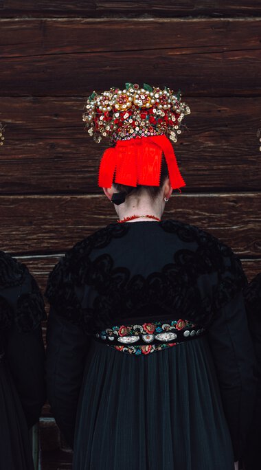 Drei Frauen in traditionellen Trachten mit bunten Kopftüchern und Schmuck stehen vor einer Holzfassade, Rückansicht. | ©  Kleinwalsertal Tourismus eGen • Fotograf: Stefan Klauser