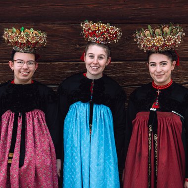 Drei junge Frauen in traditioneller Tracht mit aufwendigen Blumenkränzen auf dem Kopf, vor einer Holzwand, lachend und in Tracht gekleidet. | © Kleinwalsertal Tourismus eGen • Fotograf: Stefan Klauser