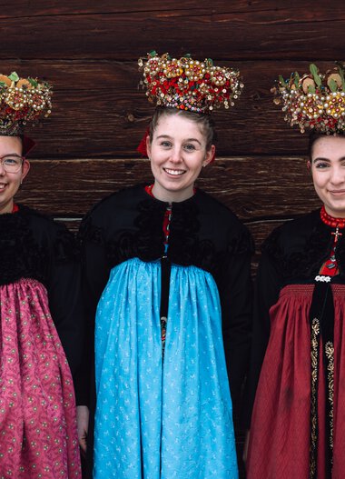 Drei junge Frauen in traditioneller Tracht mit aufwendigen Blumenkränzen auf dem Kopf, vor einer Holzwand, lachend und in Tracht gekleidet. | © Kleinwalsertal Tourismus eGen • Fotograf: Stefan Klauser