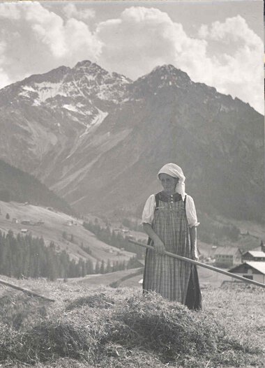 Historisches schwarz-weiß Foto von zwei Bauern in Tracht beim Heuen auf einer Wiese mit Berglandschaft im Hintergrund. | © Kleinwalsertal Tourismus eGen