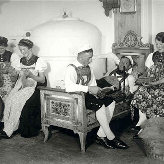 Historische Schwarz-Weiß-Aufnahme von fünf Frauen in traditionellen Trachten, die im Wohnzimmer sitzen und unterschiedliche Handarbeiten und Musikstücke machen. | © Kleinwalsertal Tourismus eGen