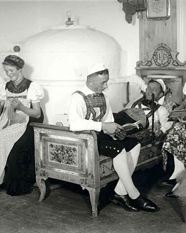 Historische Schwarz-Weiß-Aufnahme von fünf Frauen in traditionellen Trachten, die im Wohnzimmer sitzen und unterschiedliche Handarbeiten und Musikstücke machen. | © Kleinwalsertal Tourismus eGen
